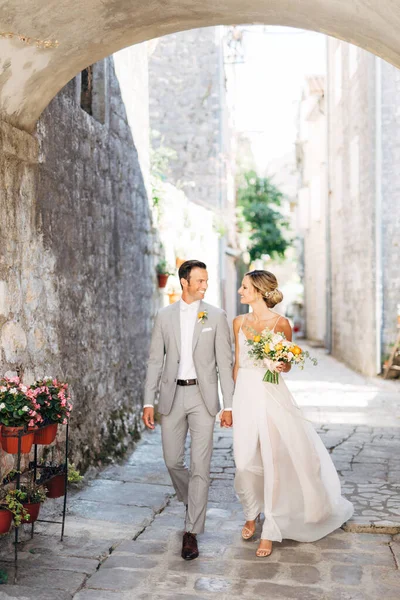 The bride and groom with a bouquet walk hand in hand along a cozy street of the old city under the arch and smile — Stock Photo, Image