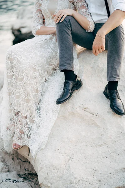 Noiva e noivo estão sentados em uma grande pedra na costa. Noiva colocar as mãos sobre os joelhos do noivo — Fotografia de Stock
