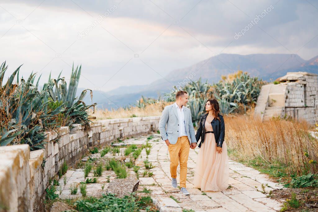 Handsome groom in a bright suit and bride in a leather jacket walk hand in hand through the ruins of an ancient castle