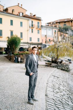 Damat elleri ceplerinde kaldırımlı bir sokakta antik villaların arka planında duruyor. Como Gölü