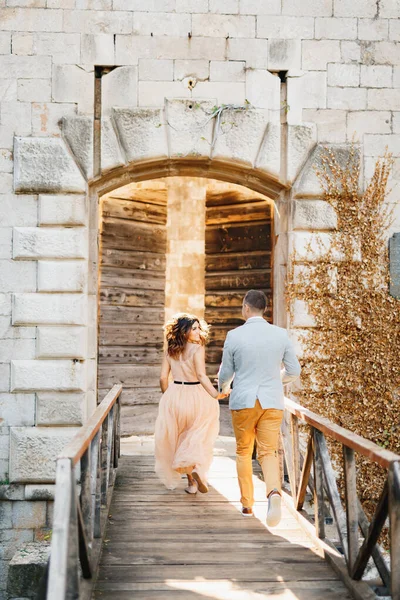 Friss házasok futnak át a hídon, kéz a kézben a kastély nyitott faajtajában. — Stock Fotó