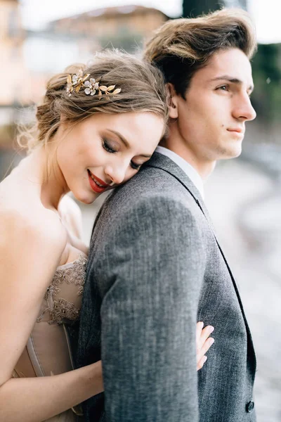 Pengantin wanita yang tersenyum memeluk pengantin pria dari belakang sambil berdiri di trotoar Danau Como. Setengah-potret — Stok Foto