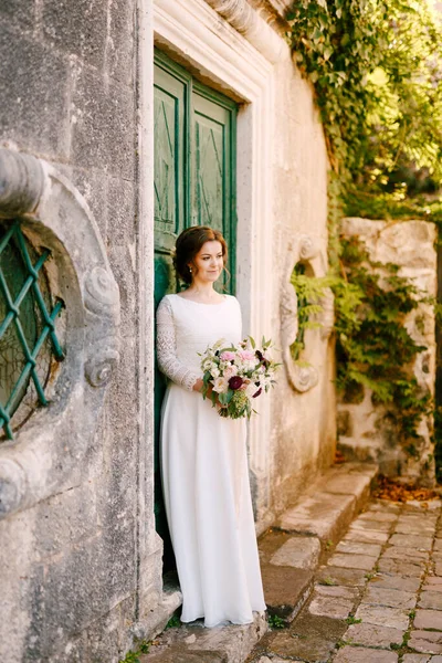 Leende bruden står mot bakgrunden av en byggnad med en vacker bukett blommor i händerna — Stockfoto