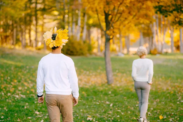 El hombre en una corona de hojas amarillas sigue a la mujer vestida con ropa ligera en el bosque de otoño. Vista trasera —  Fotos de Stock