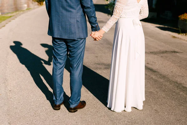 Les jeunes mariés se tiennent debout sur la route en se tenant la main. Le soleil brille brillamment dans leur dos. Des silhouettes de mariés jettent une ombre sur la route — Photo