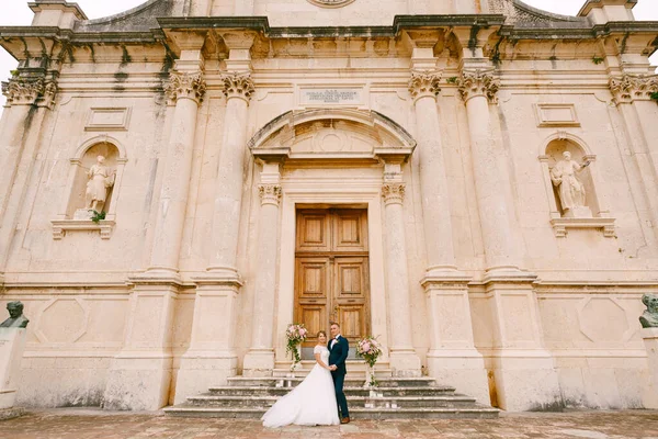 Bruden och brudgummen står kramas nära dörren på trappan till Födelsekyrkan av Jungfru Maria kyrka i Prcanj — Stockfoto