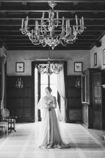 Pengantin dalam gaun yang indah dengan kerudung dan karangan bunga berdiri di tengah ruangan. Danau Como. Foto hitam dan putih — Stok Foto