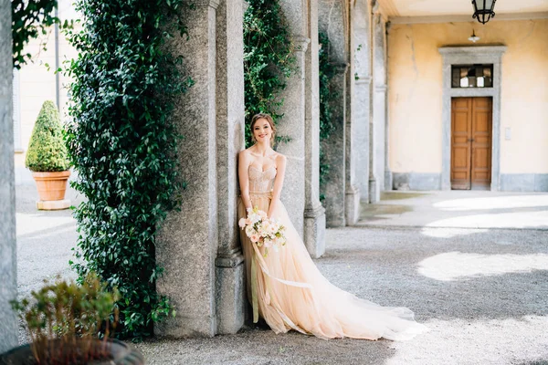 Leende brud i en klänning med en bukett rosa blommor lutande mot en pelare i ett välvt rum. Comosjön, Italien — Stockfoto