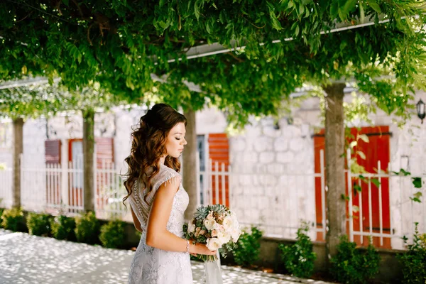 Gyönyörű menyasszony fényűző ruhában áll egy gyönyörű esküvői csokor rózsával egy zöld kertben egy napsütéses napon.. — Stock Fotó