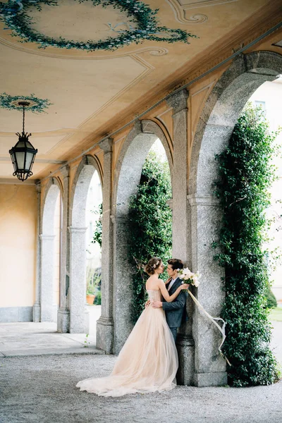 Novia con un ramo de flores abraza novio apoyado en un arco entrelazado con hiedra verde. Lago de Como —  Fotos de Stock