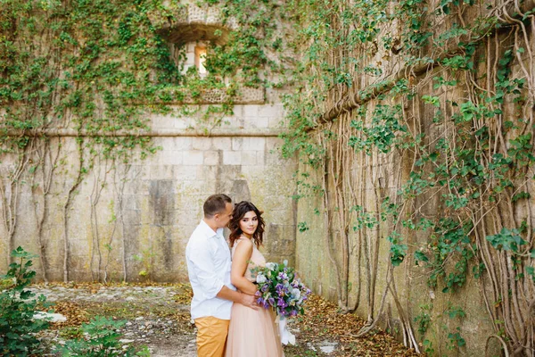 Sposo abbraccia sposa in un bellissimo abito pastello con mazzo di fiori sullo sfondo di un muro di pietra, intrecciato con edera — Foto Stock