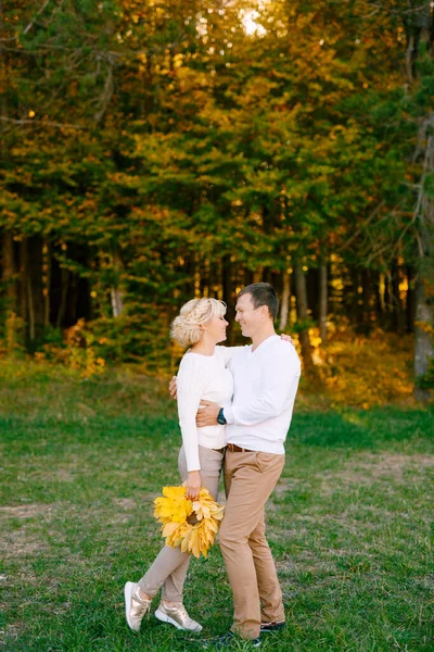 Muž v lehkém oblečení s úsměvem objímá ženu s kyticí žlutých listů v podzimním lese — Stock fotografie
