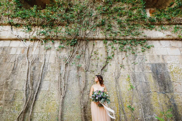 Bella sposa in un abito pastello con un lussuoso mazzo di fiori si erge sullo sfondo di un muro di pietra intrecciato con edera verde — Foto Stock