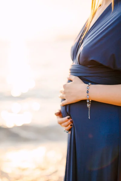Güzel bir manikürle uzun mavi elbiseli hamile kadın elleriyle karnına sarılıyor. — Stok fotoğraf