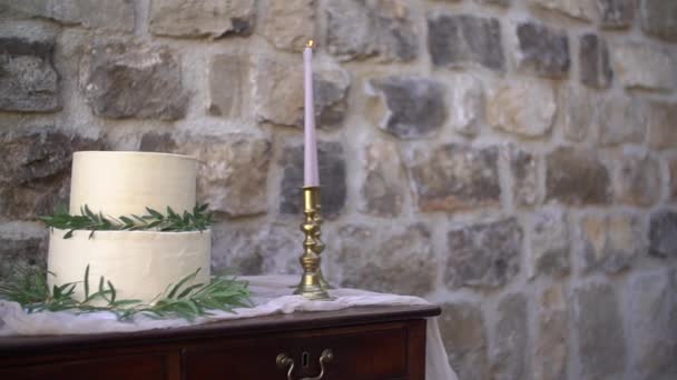 Bílý krémový dort, zdobený olivovými a eukalyptovými sprigy, vedle hořících svíček ve zlatých svícnech na dřevěném stole se zásuvkami — Stock video