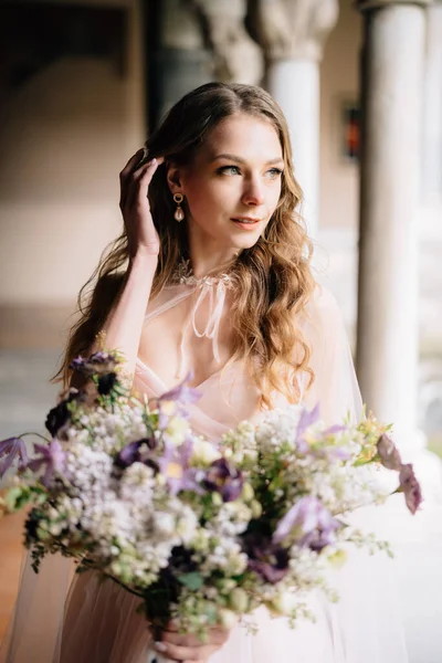 Bruden i en vacker rosa klänning håller en bukett vilda blommor i handen och stoppar en hårlock bakom örat mot bakgrund av en gammal byggnad — Stockfoto