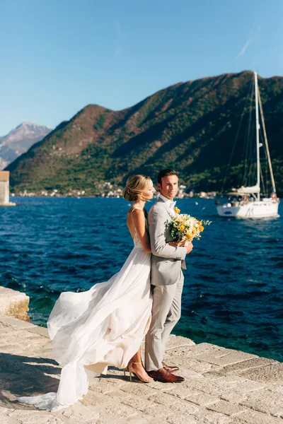 A menyasszony és a vőlegény ölelkeznek a mólón a Kotori-öbölben, mögöttük zöld hegyek és egy fehér jacht. — Stock Fotó