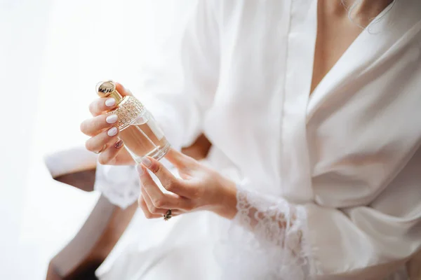 Sposa in abito di pizzo bianco seduta su una poltrona tiene in mano una bottiglia di profumo — Foto Stock