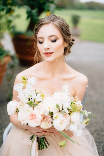 Bruid sloot haar ogen in een prachtige jurk met een boeket bloemen tegen de achtergrond van een groene boom in een bad. Comomeer — Stockfoto