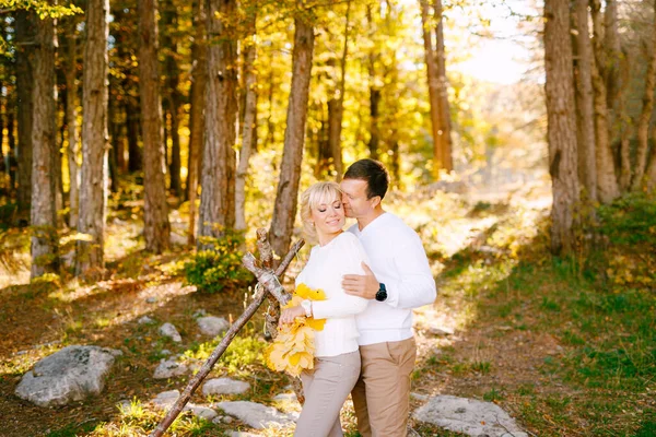 Мужчина целует и обнимает улыбающуюся женщину за плечи. У женщины в руках жёлтые листья. Широкий угол — стоковое фото