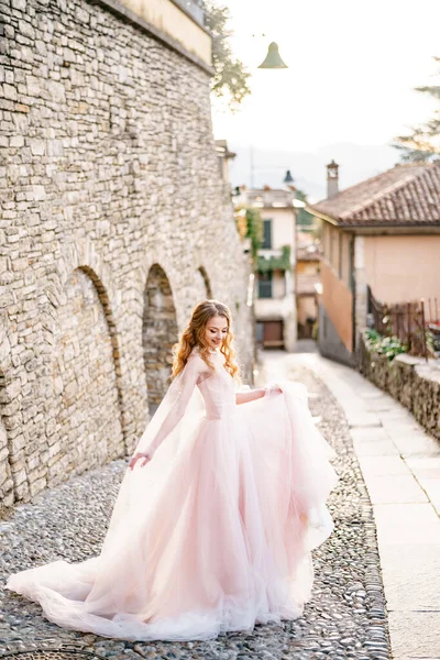 Novia sonriente en un hermoso vestido largo de color rosa se encuentra en los adoquines de una antigua calle de Bérgamo, Italia —  Fotos de Stock