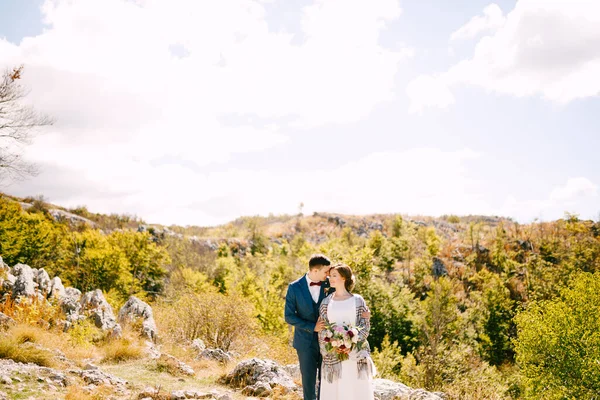Ο γαμπρός αγκαλιάζει τη νύφη με ένα καρό σάλι με ένα μπουκέτο λουλούδια στα χέρια της στο φόντο βράχων και πράσινων φυτών. — Φωτογραφία Αρχείου