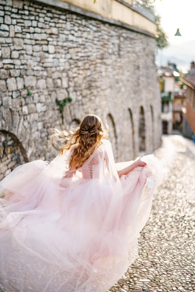 Novia con un vestido revoloteando en el viento corre a lo largo de los adoquines de una antigua calle en Bérgamo, Italia. Vista trasera —  Fotos de Stock