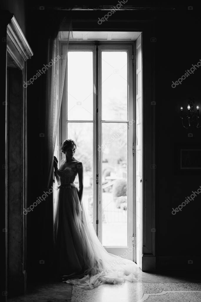 Bride is standing next to a large window, turning sideways. Lake Como. Black and white photo
