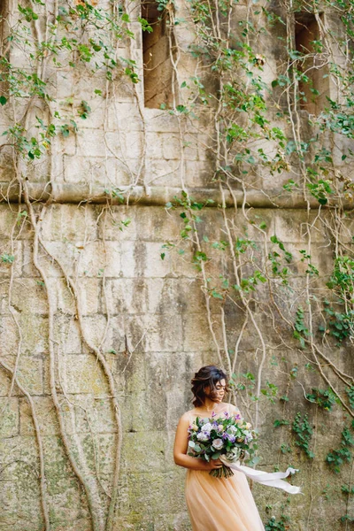 Gyönyörű menyasszony egy krém esküvői ruha egy csokor virágot a háttérben egy kőfal a Mamula sziget Montenegróban — Stock Fotó