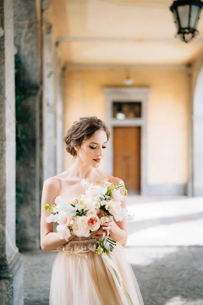 Bruid in een prachtige jurk met een boeket van roze bloemen staat in een boogvormige gang. Comomeer, Italië — Stockfoto