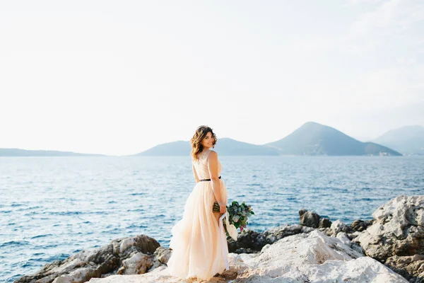 Linda noiva em um vestido de noiva pastel fica sorrindo em uma rocha acima do mar com um buquê de flores em suas mãos — Fotografia de Stock