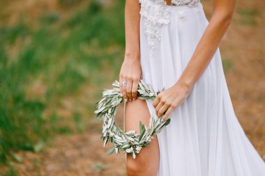 Beyaz işlemeli elbiseli güzel gelin bir zeytin yaprağı çelengi tutuyor. Gelin bacağı elbisenin kesiğinden görülebiliyor.