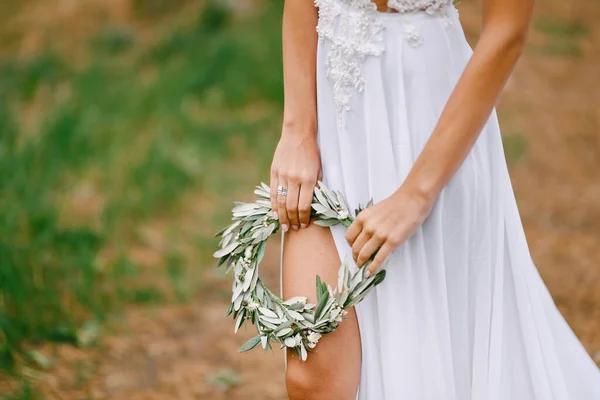 Beyaz işlemeli elbiseli güzel gelin bir zeytin yaprağı çelengi tutuyor. Gelin bacağı elbisenin kesiğinden görülebiliyor. — Stok fotoğraf