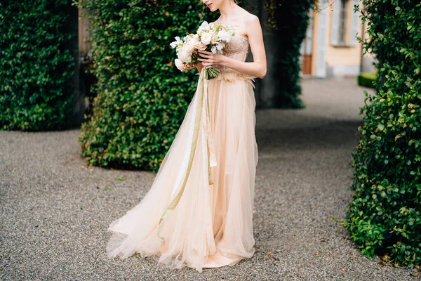 Bruden i en rosa klänning håller en bukett blommor mot bakgrund av en byggnad sammanflätad med grönska. Comosjön, Italien — Stockfoto