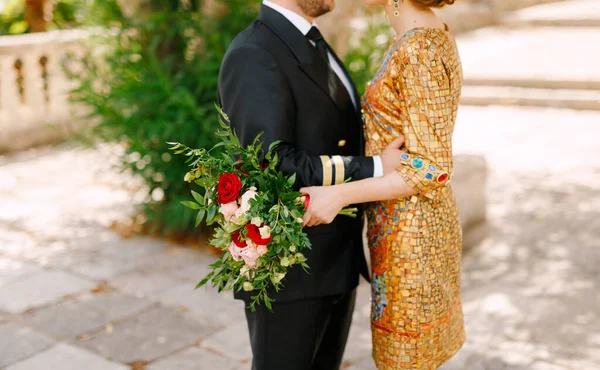 Η νύφη σε ένα ασυνήθιστο χρυσό φόρεμα και ο γαμπρός σε ένα κοστούμι αγκαλιάζουν, η νύφη κρατώντας μπουκέτο στα χέρια της — Φωτογραφία Αρχείου