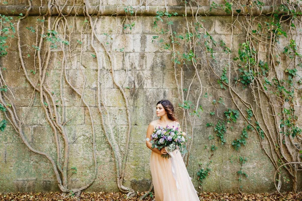 Gyönyörű menyasszony bézs menyasszonyi ruhában, gyönyörű virágcsokorral a hátterében egy borostyánnal összefonódó kőfallal. — Stock Fotó