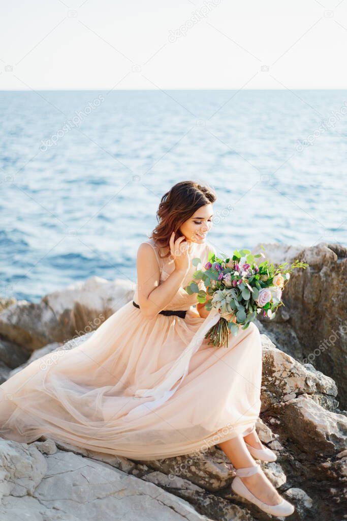 Beautiful smiling bride in a beige dress with a bouquet of flowers in her hands sits on a rock above the sea