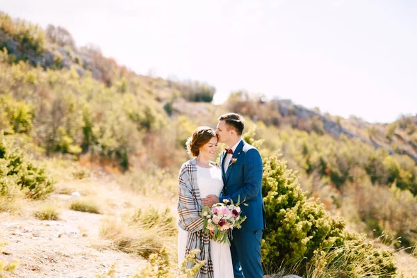 Groom embrasser mariée dans un châle à carreaux avec un bouquet de fleurs dans ses mains sur le fond de roches et de plantes vertes — Photo