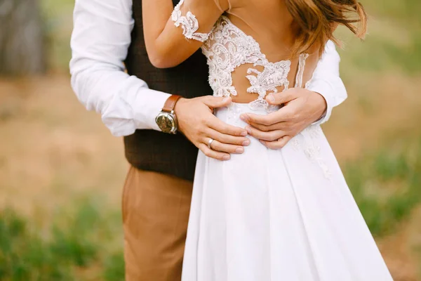 Bräutigam hält seine Hände auf der Taille der Braut in einem schönen bestickten weißen Kleid. Braut steht mit umgedrehtem Rücken — Stockfoto