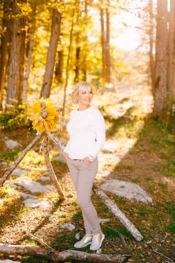 Beyaz kazaklı gülümseyen kadın bir ağacın yanında duruyor. Sarı yapraklardan bir çelenk dallarda asılı.