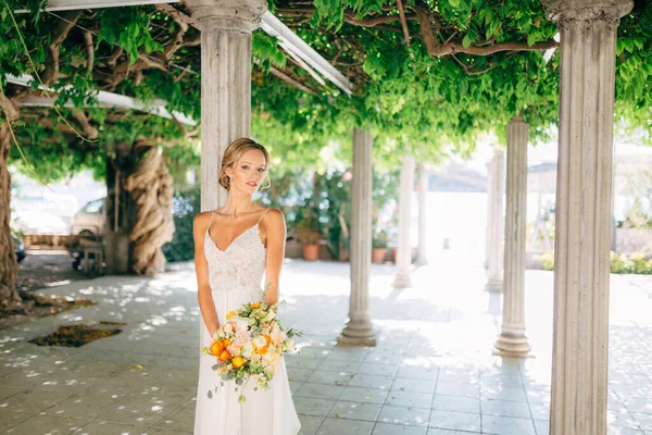 Pengantin wanita berdiri di antara kolom putih dikepang dengan tanaman merambat hijau dan memegang karangan bunga pernikahan dengan buttercups oranye — Stok Foto