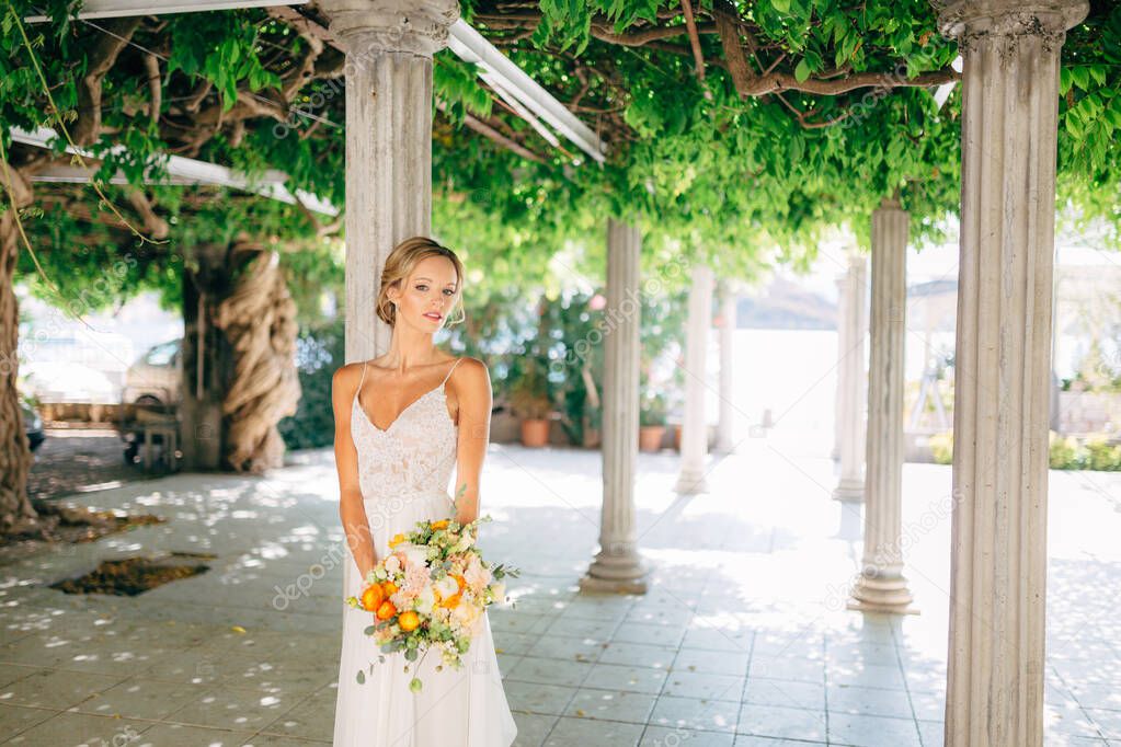 The bride stands among white columns braided with green vines and holds a wedding bouquet with orange buttercups 
