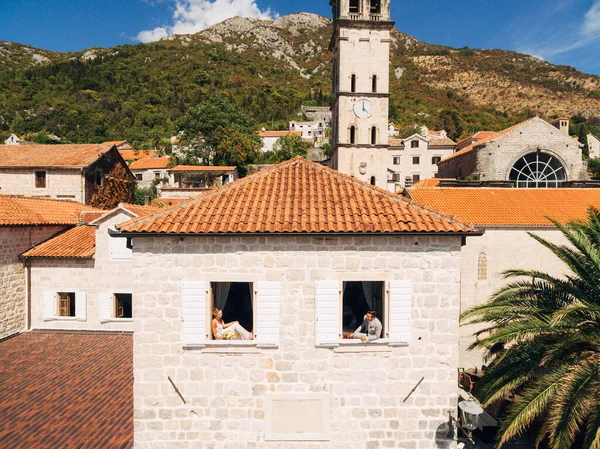 Жених и невеста выглядывают из окон соседнего дома с оранжевой крышей в старом городе Пераст — стоковое фото