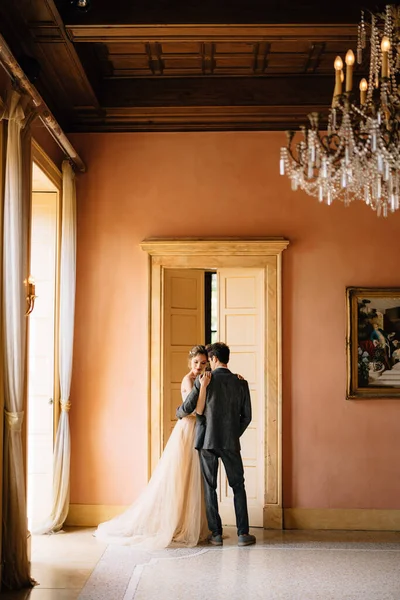 Bruid omhelst bruidegom tegen de achtergrond van de hoge deur naar de kamer van de oude villa. Comomeer — Stockfoto