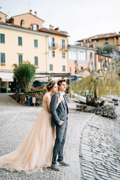 Bride hugs from behind groom while standing on the embankment of Lake Como against the background of old houses — Stock Photo, Image