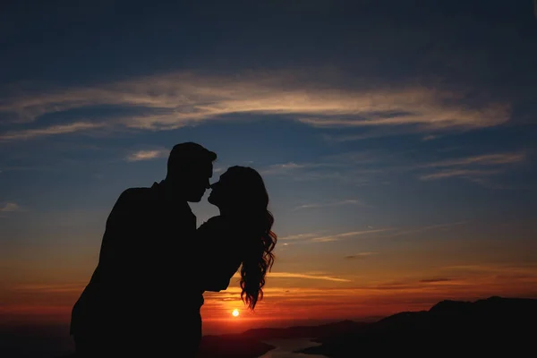 Silhouetten van de bruid en bruidegom die elkaar omhelzen en kussen op de berg Lovcen met uitzicht op de baai van Kotor bij zonsondergang — Stockfoto