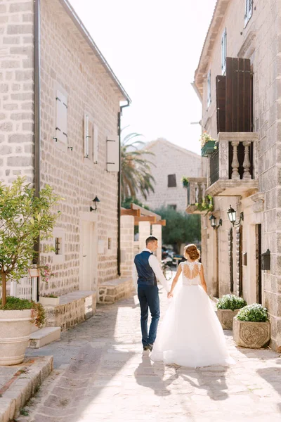 Les mariés marchent le long d'une rue étroite et confortable de la vieille ville de Perast en se tenant la main, vue arrière — Photo