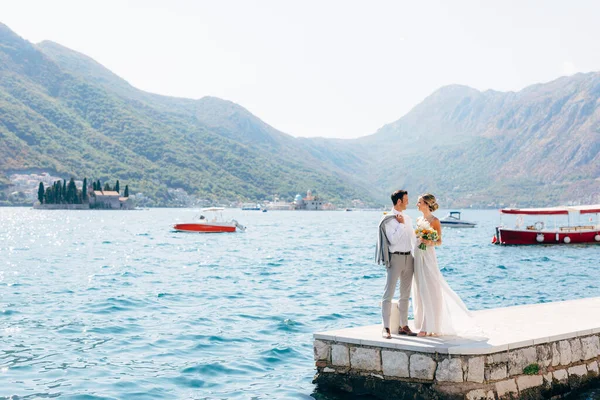 Η νύφη και ο γαμπρός στέκονται δίπλα-δίπλα στην προβλήτα στον κόλπο του Κότορ, τα νησιά Πέραστ είναι πίσω τους. — Φωτογραφία Αρχείου