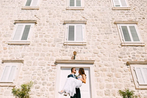 Groom holding bride in his arms against the stone wall of the building on a bright sunny day. — Stock Photo, Image