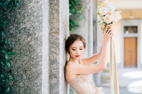 La novia bajó los ojos, apoyándose en un pilar con las manos levantadas con un ramo de flores. Lago de Como —  Fotos de Stock